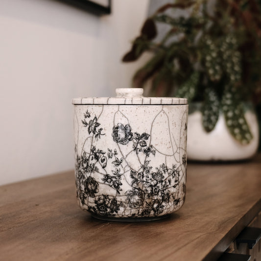 Ceramic candle holder with black floral pattern on a wooden surface with a plant in the background